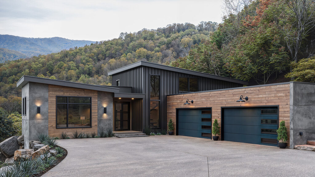 Modern house with a mix of wood, concrete, and metal exterior, flat and sloped roofs, large glass door and windows, two blue garage doors, surrounded by trees and hills in the background.