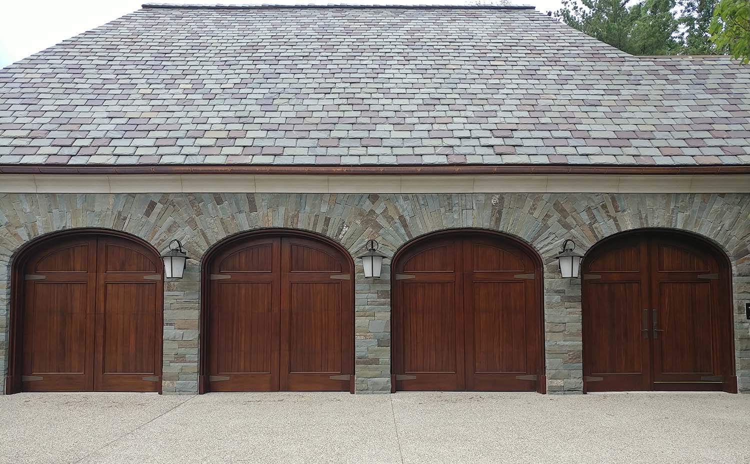 A four-car garage features wooden doors with arched tops set into a stone exterior wall. Each of the three central doors has a light fixture above it, while the fourth door on the far right includes a double door entrance for walking. The roof is made of slate shingles.